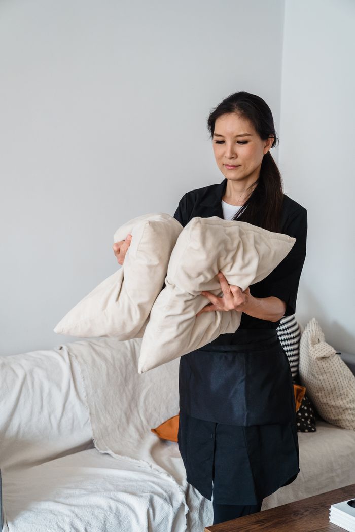 home-img Smiling housekeeper arranging pillows on a sofa in a bright living room.